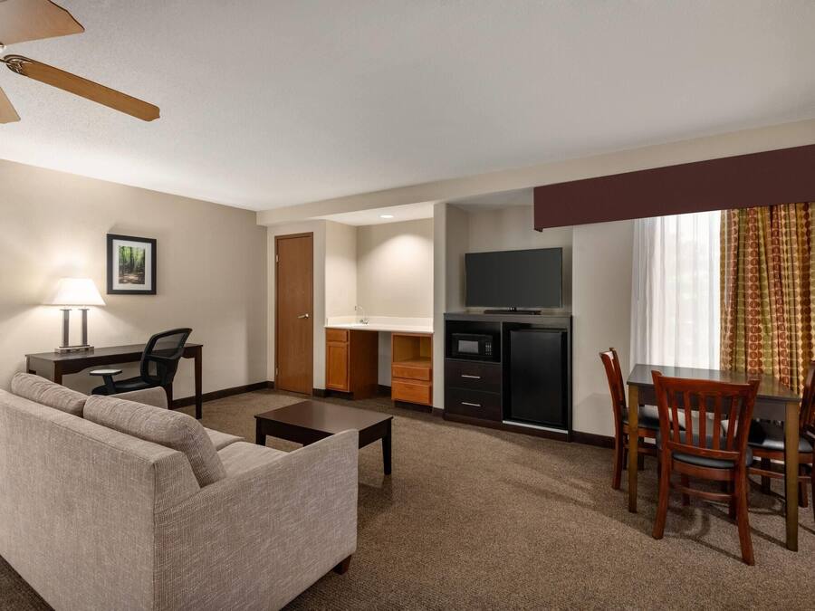 A hotel room with a sofa, a dining table with chairs, a kitchenette, a TV, and a ceiling fan.