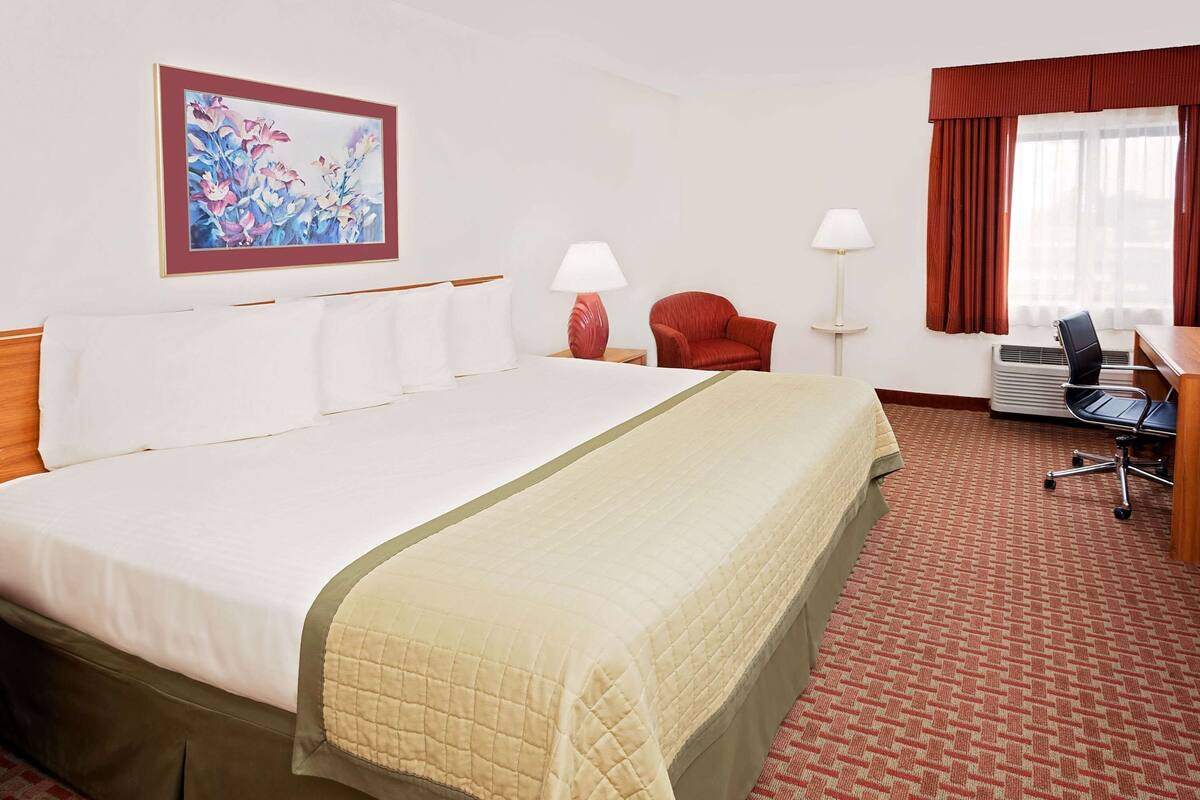 A hotel room with a large bed, a desk with a chair, a red armchair, a painting on the wall, and a window with curtains.