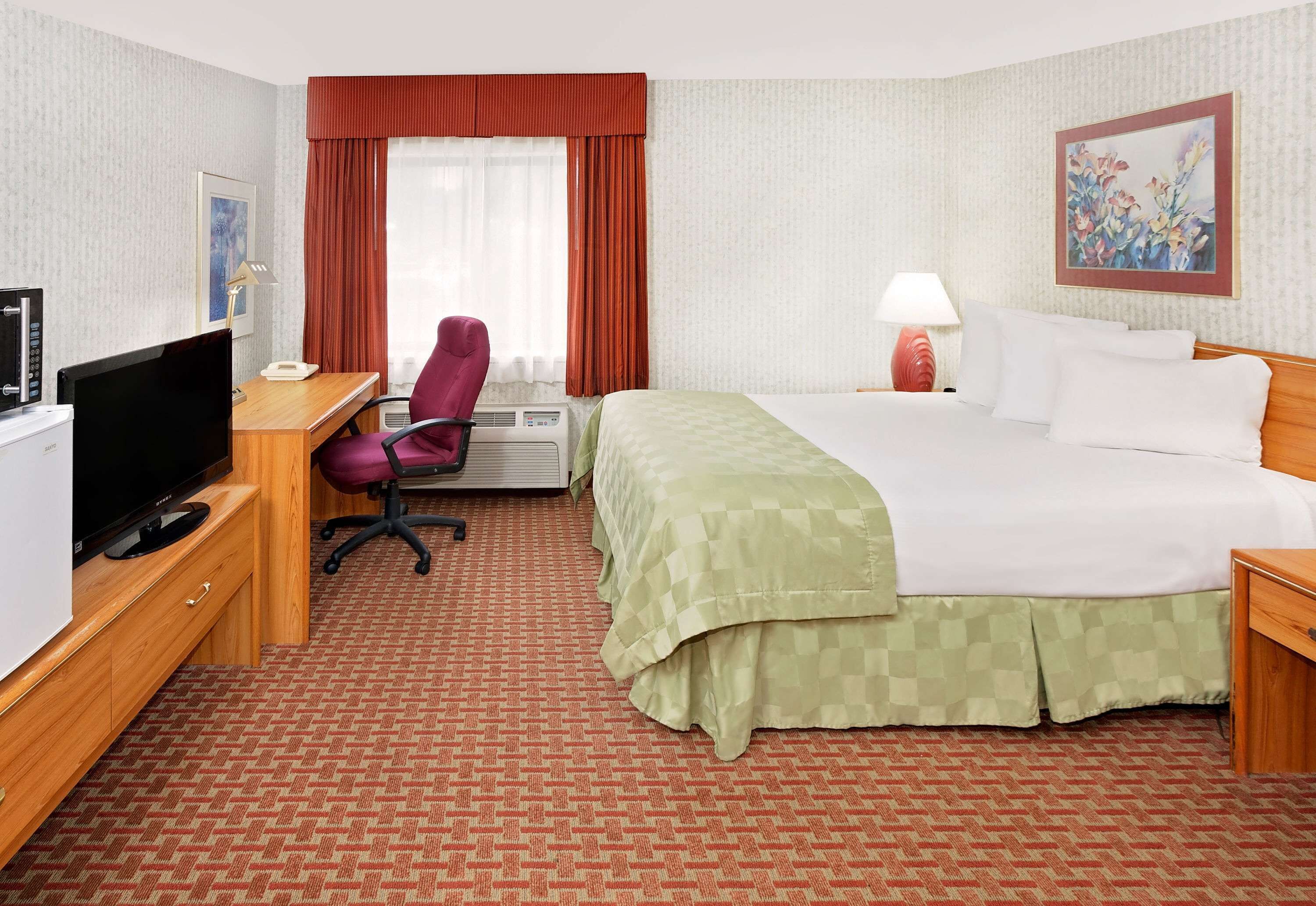 Habitación de hotel con cama, escritorio con televisor, una silla, lámpara y un cuadro en la pared.