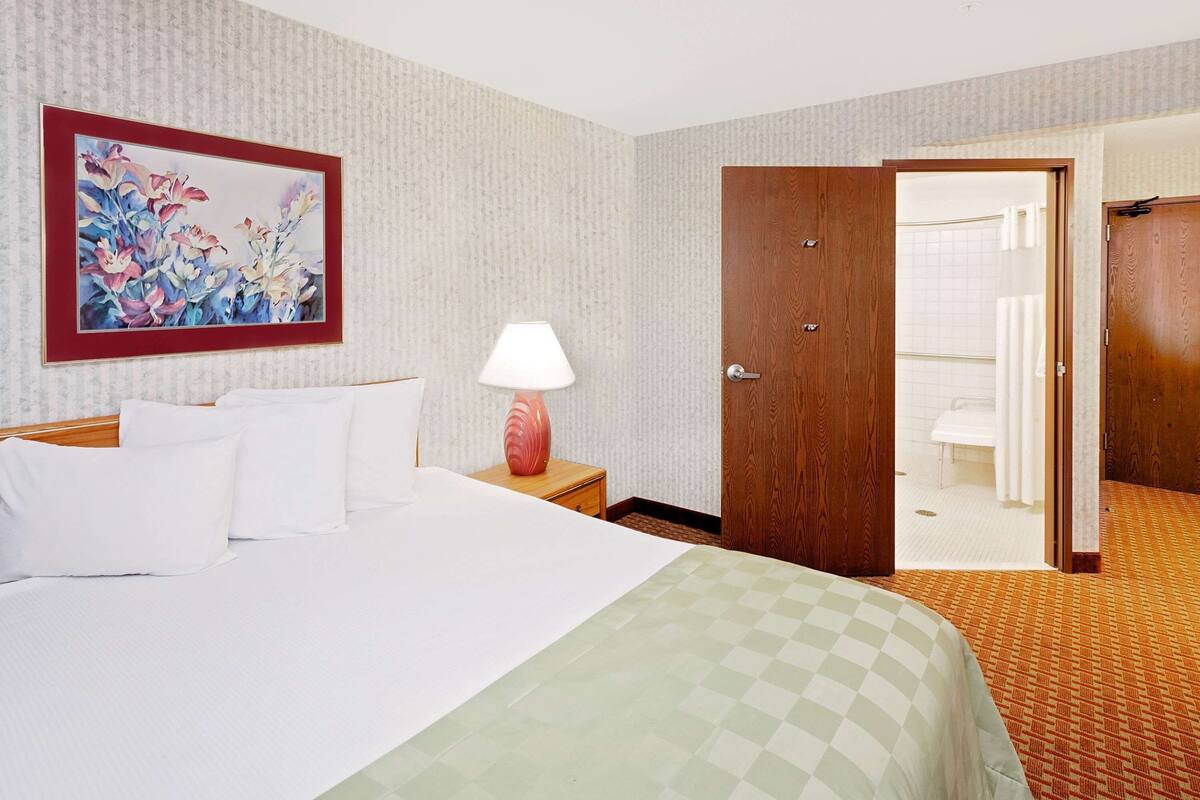 A hotel room with a bed, a nightstand with a lamp, a wooden door, and a bathroom visible through an open door.