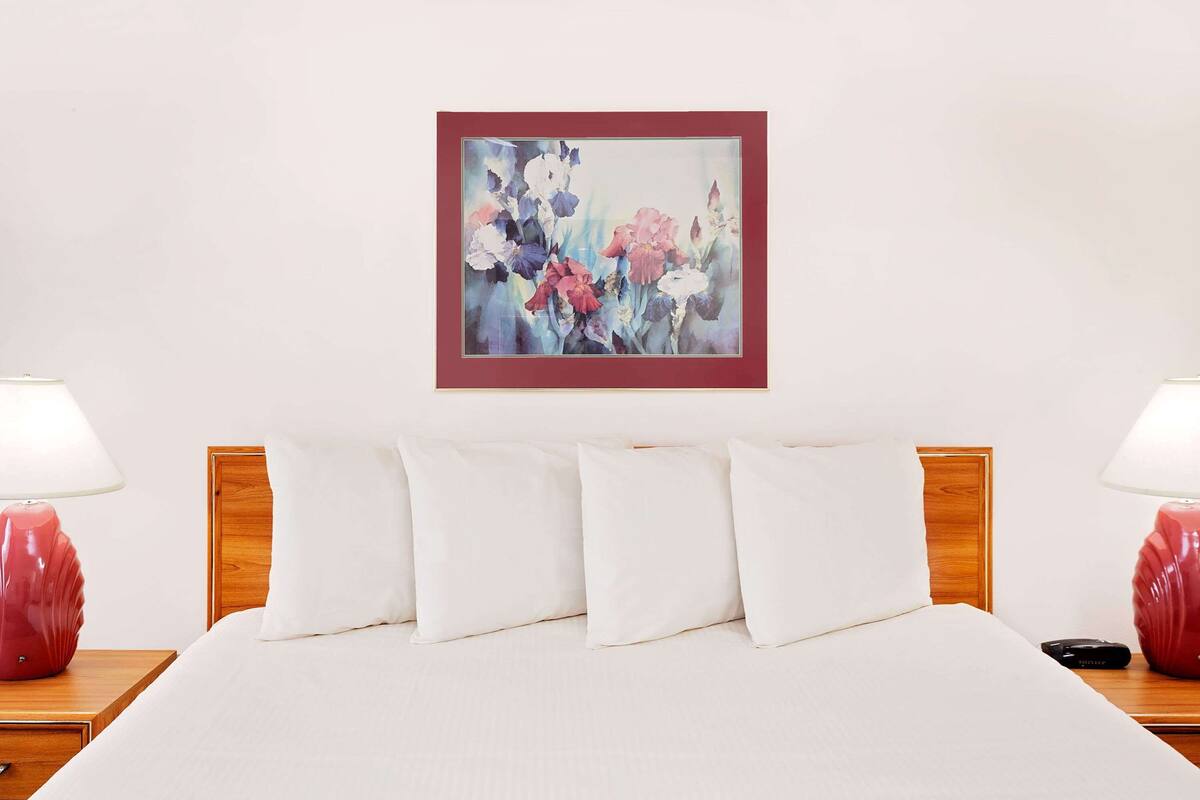 A neatly made bed with white bedding, a wooden headboard, and a framed floral artwork on the wall.