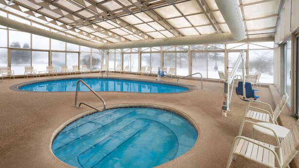 Una piscina interior con paredes de vidrio transparente, varias sillas de descanso y una escalera visible.