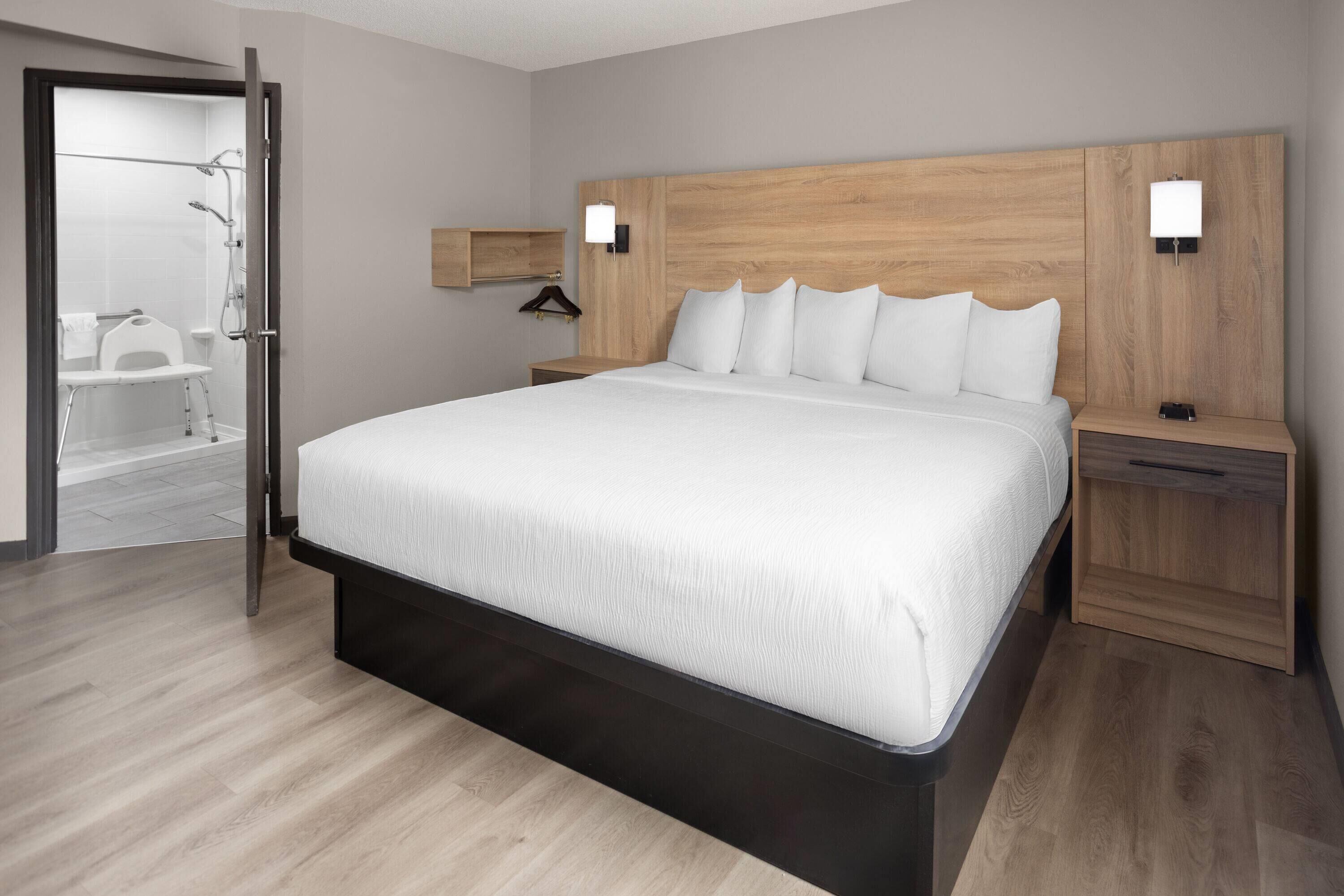 Moderni hotellihuone, jossa on suuri sänky, puinen pääty, yöpöydät ja erillinen suihkutila.