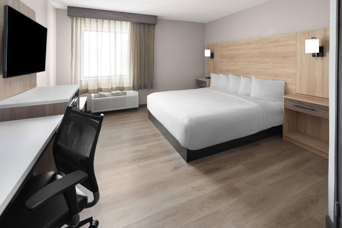 Ein Hotelzimmer mit einem Bett, einem Schreibtisch mit Stuhl, einem an der Wand befestigten Fernseher und einem Fenster mit Vorhängen.