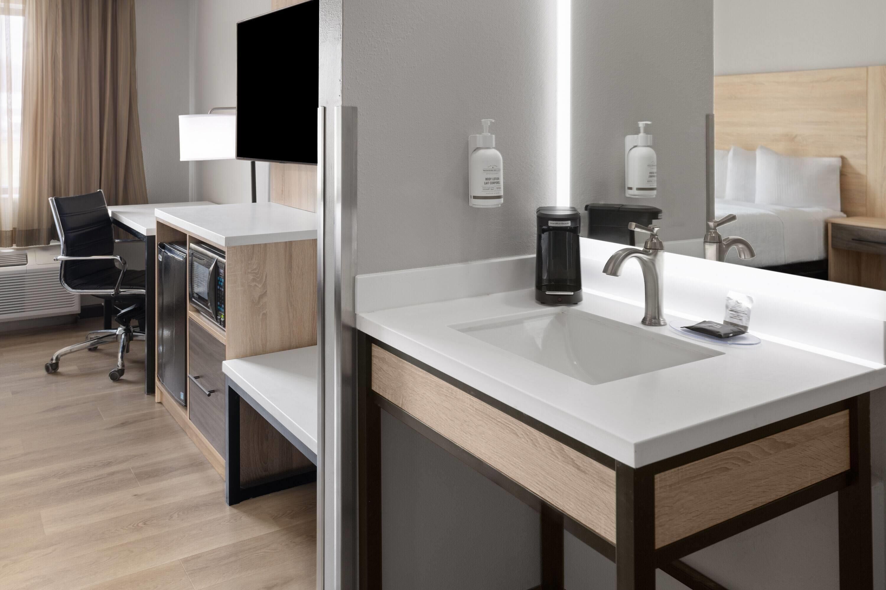 A modern hotel room with a large white sink, a flat-screen TV mounted on the wall, a desk with a chair, and a bed with white linens.