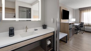 Una habitación de hotel moderna con un espejo grande, un lavamanos blanco, una cafetera, un refrigerador, un escritorio con una silla y un ventanal con cortinas.