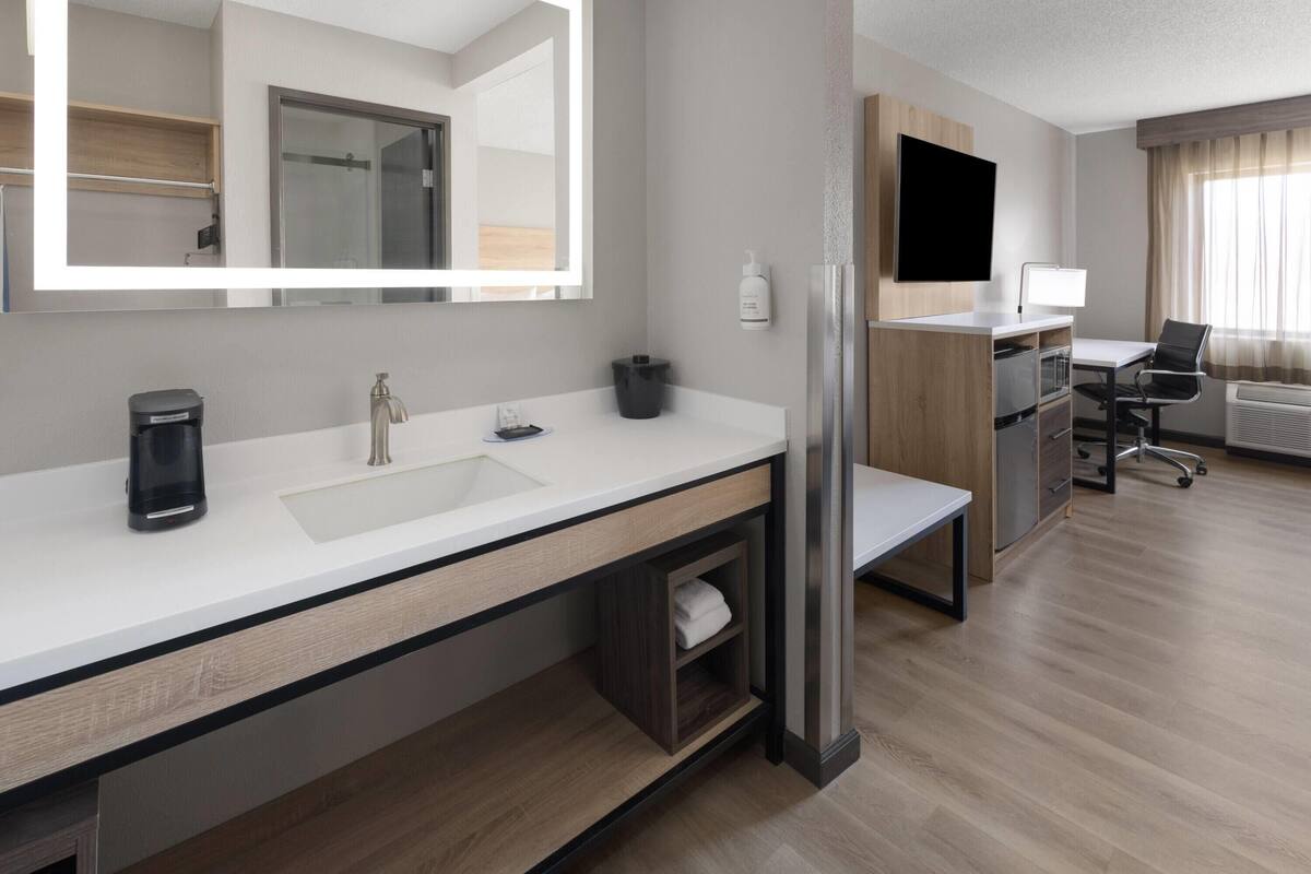 Ein modernes Hotelzimmer mit einem großen Spiegel, einem weißen Waschbecken, einer Kaffeemaschine, einem Kühlschrank, einem Schreibtisch mit Stuhl und einem großen Fenster mit Vorhängen.