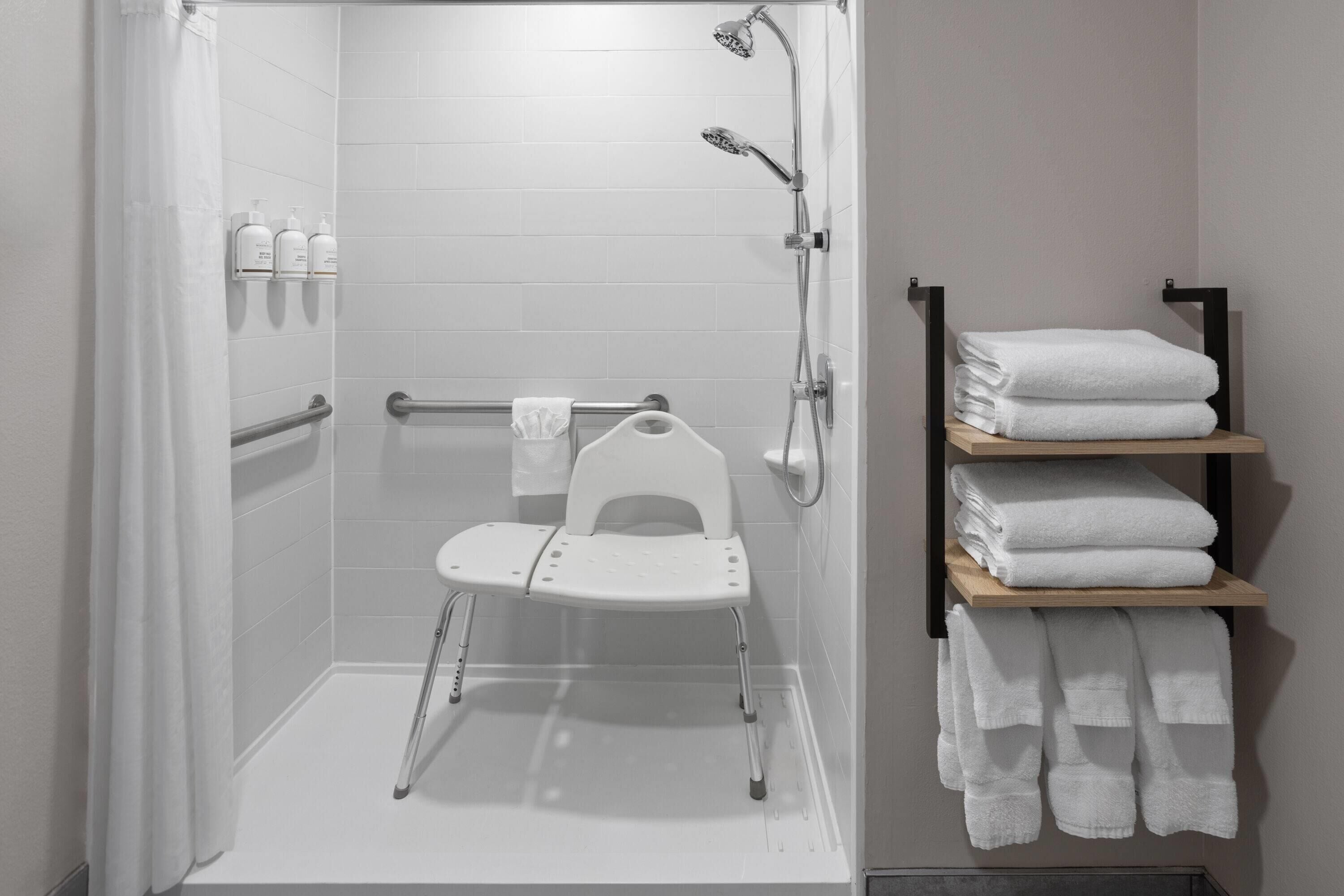 A shower area with a white chair, grab bars, and a shelf with folded towels.