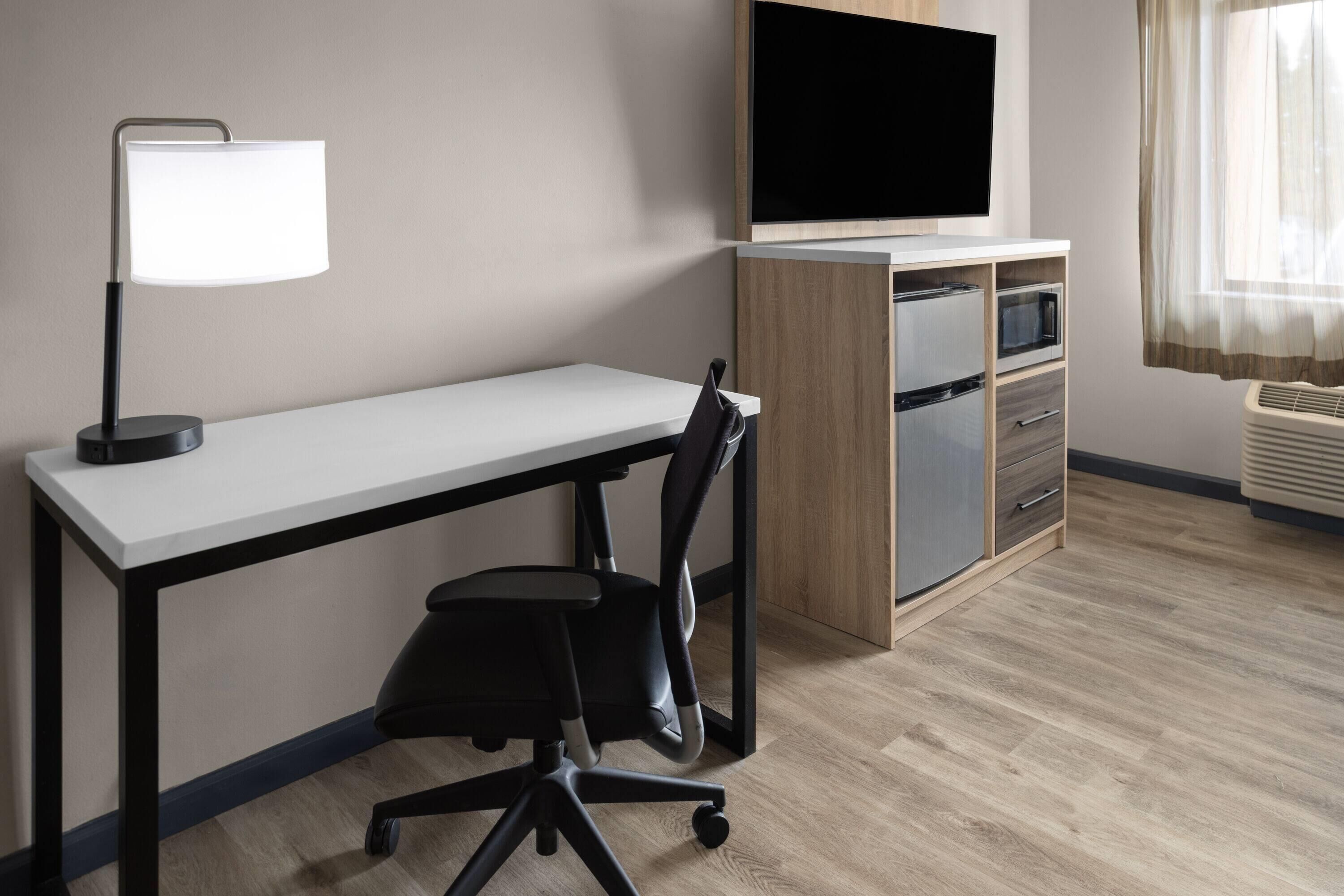 A modern hotel room with a desk, chair, TV, and built-in mini-fridge.
