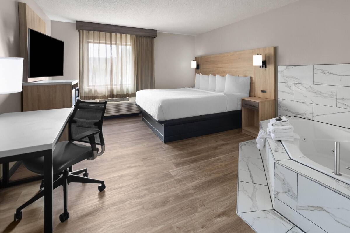 Ein Hotelzimmer mit einem Bett, einem Schreibtisch mit Stuhl, einem Fernseher, einem Badezimmer mit Badewanne und einem Fenster mit Vorhängen.