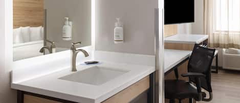 A modern hotel room with a large mirror, a white sink, a wooden desk, a black office chair, and a flat-screen TV mounted on the wall.