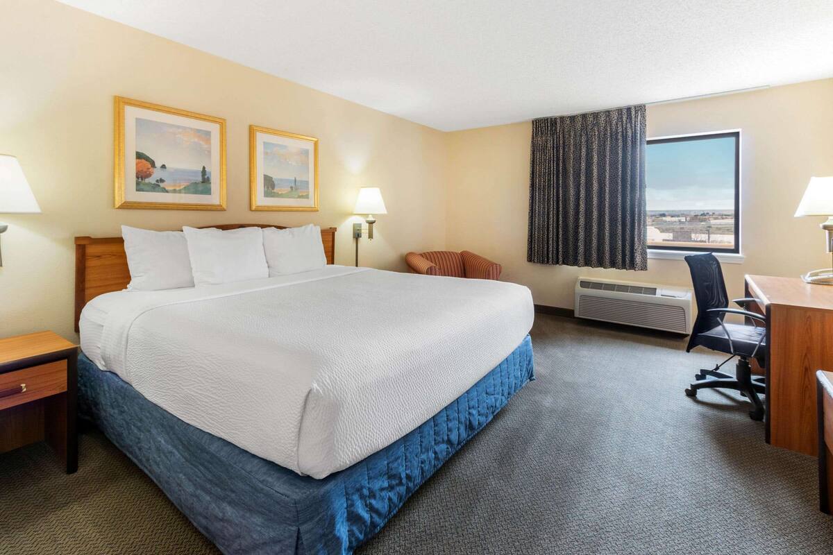 Ein Hotelzimmer mit einem großen Bett, zwei Nachttischlampen, einem Schreibtisch mit Stuhl, einem Fenster mit Aussicht und zwei gerahmten Bildern an der Wand.