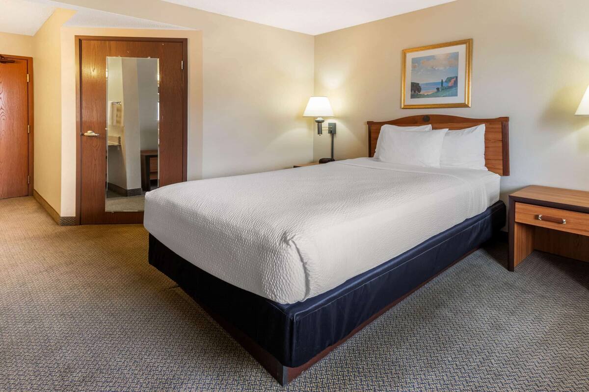 Ein Hotelzimmer mit einem großen Bett, einem Holznachttisch, einer Wandlampe und einer Tür zu einem weiteren Raum.