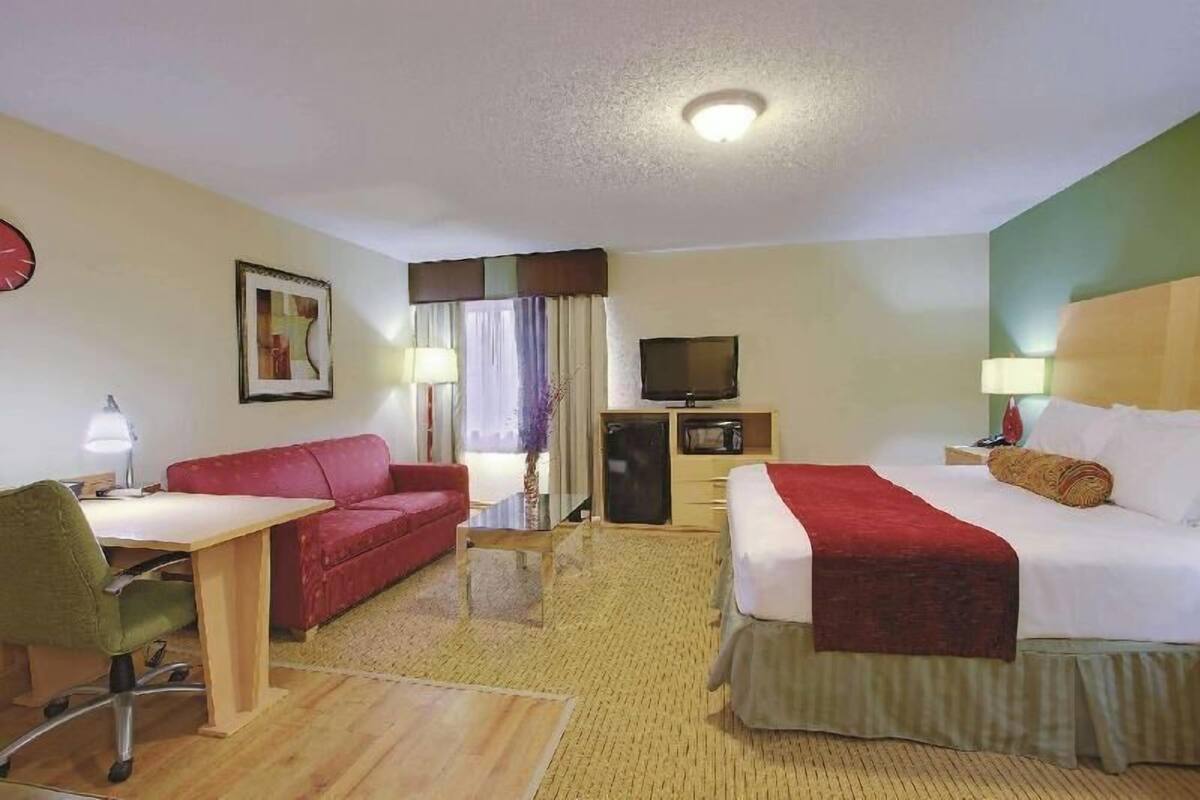 A hotel room with a bed, a desk, a red sofa, a television, and a clock on the wall.