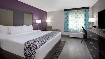 A hotel room with a large bed, a desk with a chair, a TV, and a window with patterned curtains.