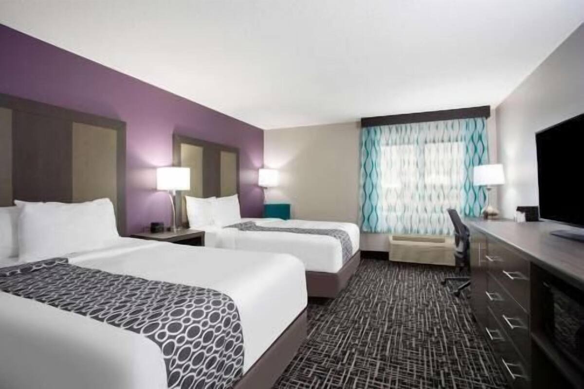 A hotel room with two beds, a desk, a TV, and a window with curtains.