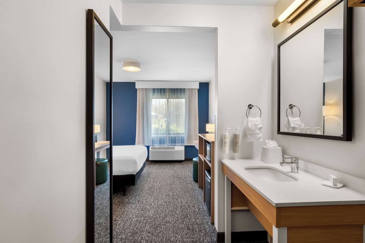 A hotel room with a bed, a desk, a bathroom with a sink and mirror, and a window with curtains.