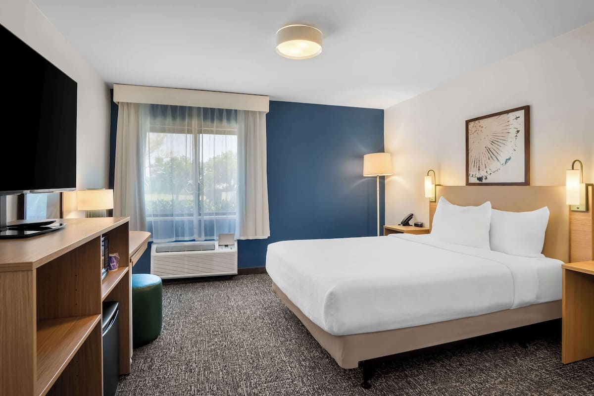 A hotel room with a bed, a desk with a TV, a lamp, a picture on the wall, and a window with curtains.