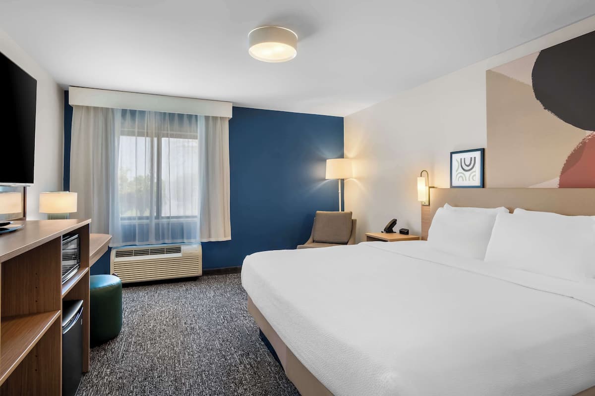 A hotel room with a large bed, a desk with a lamp, a chair, a nightstand, and a window with curtains.