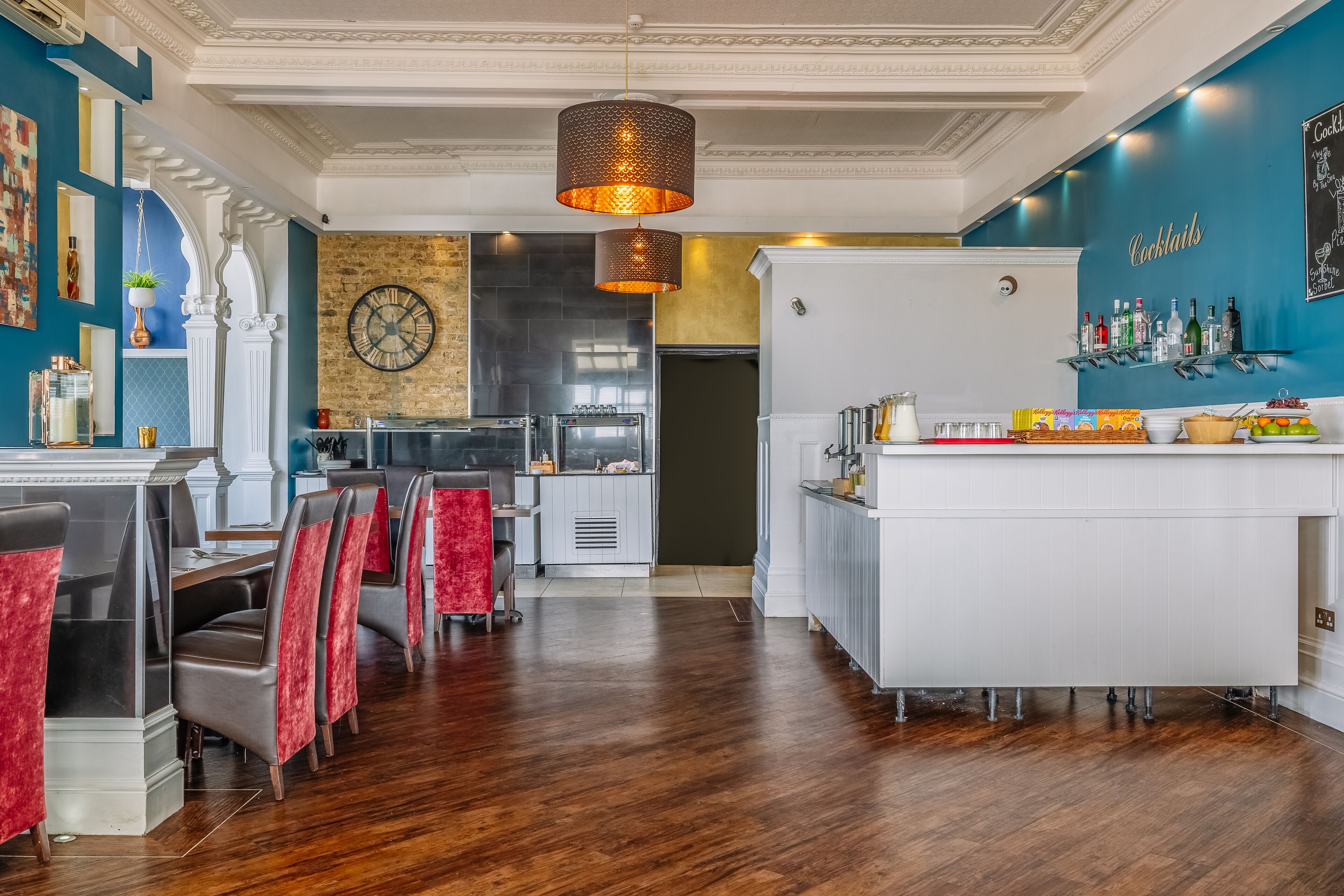 A modern restaurant interior with a bar area, wooden flooring, and red upholstered chairs.