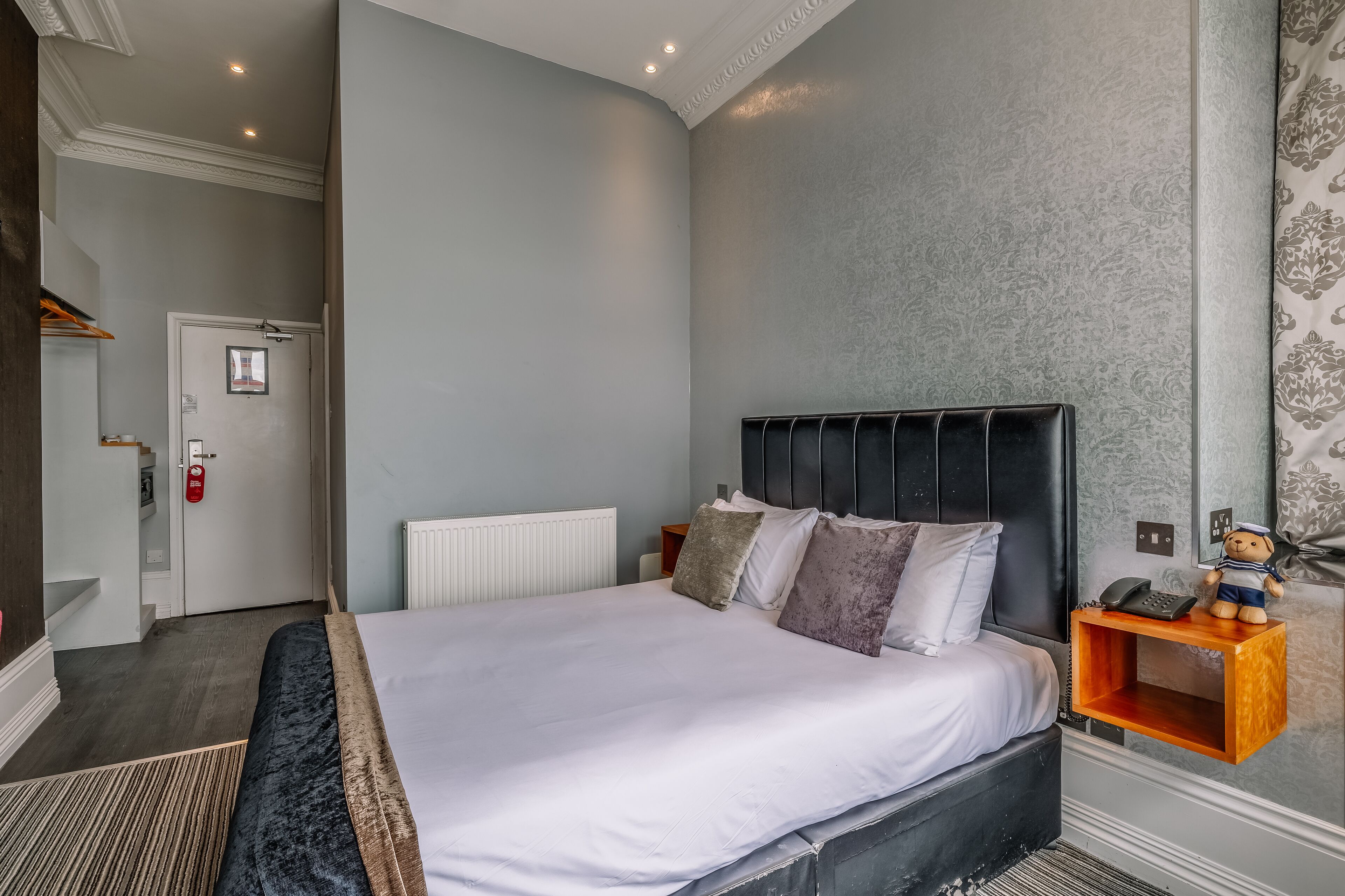 A hotel room with a large bed, a bedside table with a telephone, and a small shelf with a teddy bear.