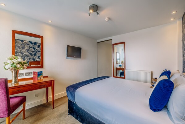 Una habitación de hotel con cama, escritorio, silla, televisor y espejo.