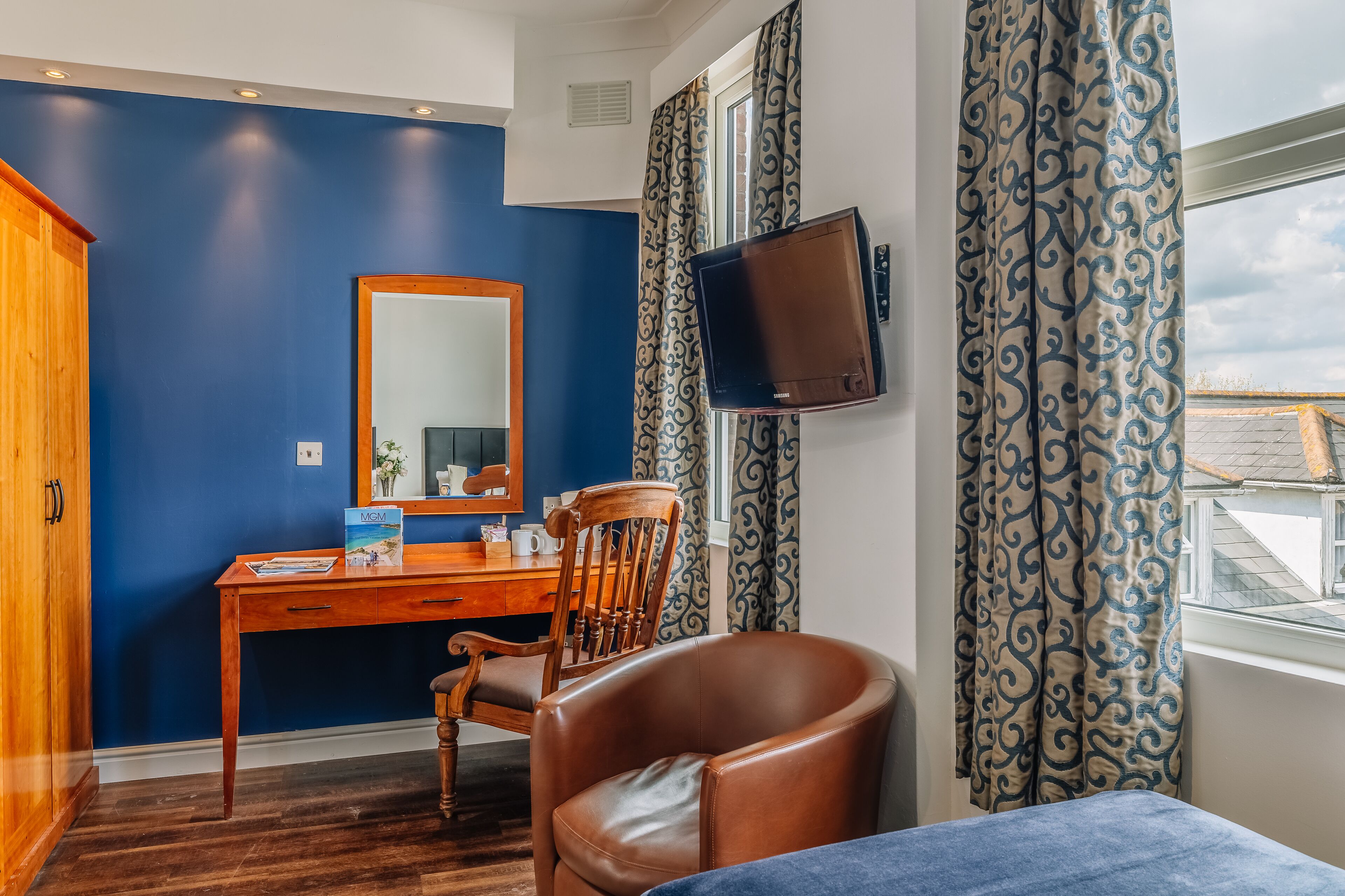 Ein Zimmer mit einem Holztisch, einem Stuhl, einem an der Wand montierten Fernseher und einem Fenster mit gemusterten Vorhängen.