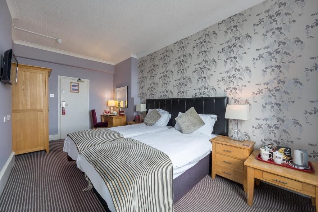 A hotel room with two beds, a wooden wardrobe, a desk with a lamp, and a floral wallpaper.