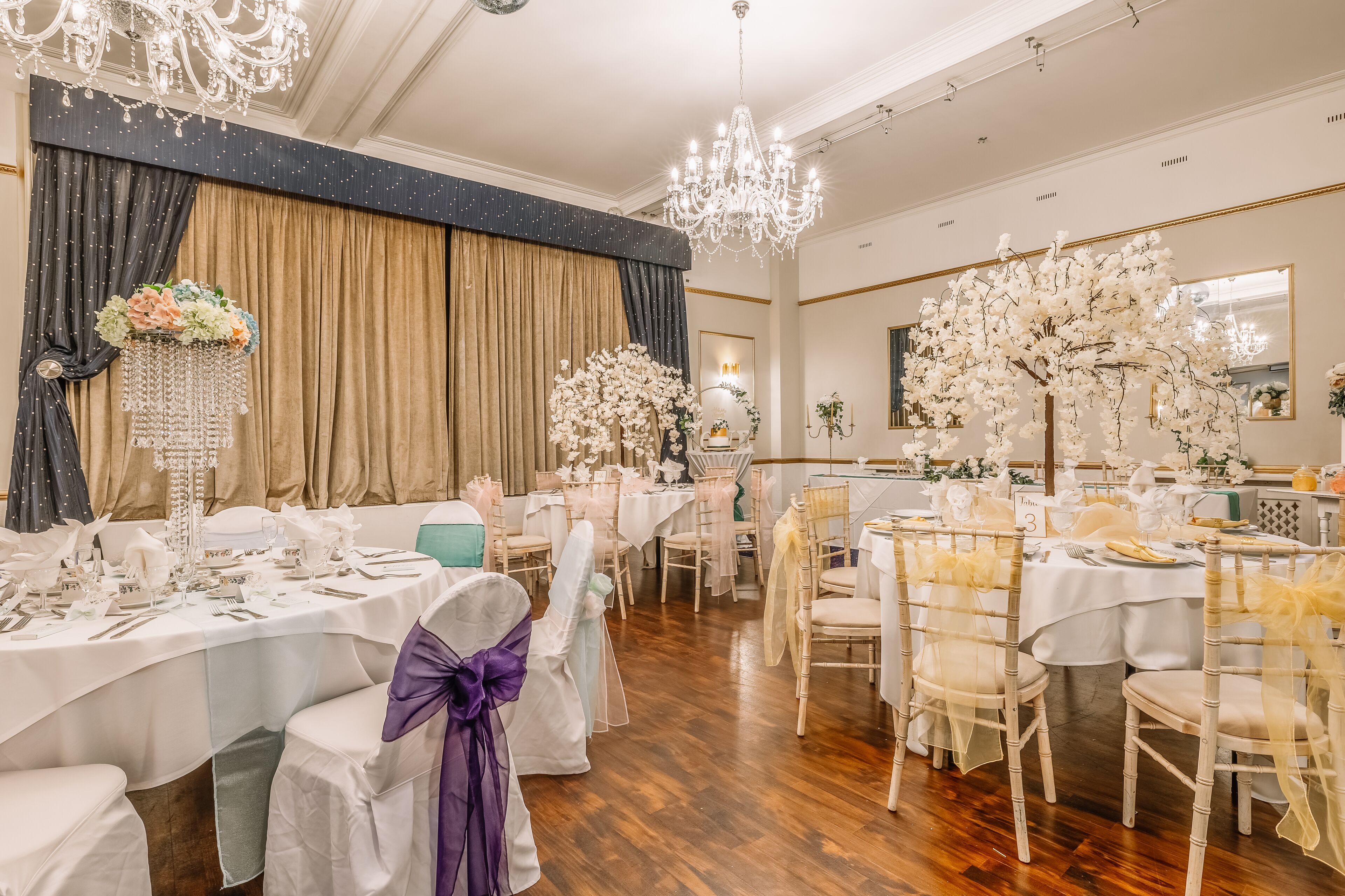 Een balzaal met ronde tafels, witte tafellakens en stoelen met witte en gouden linten.