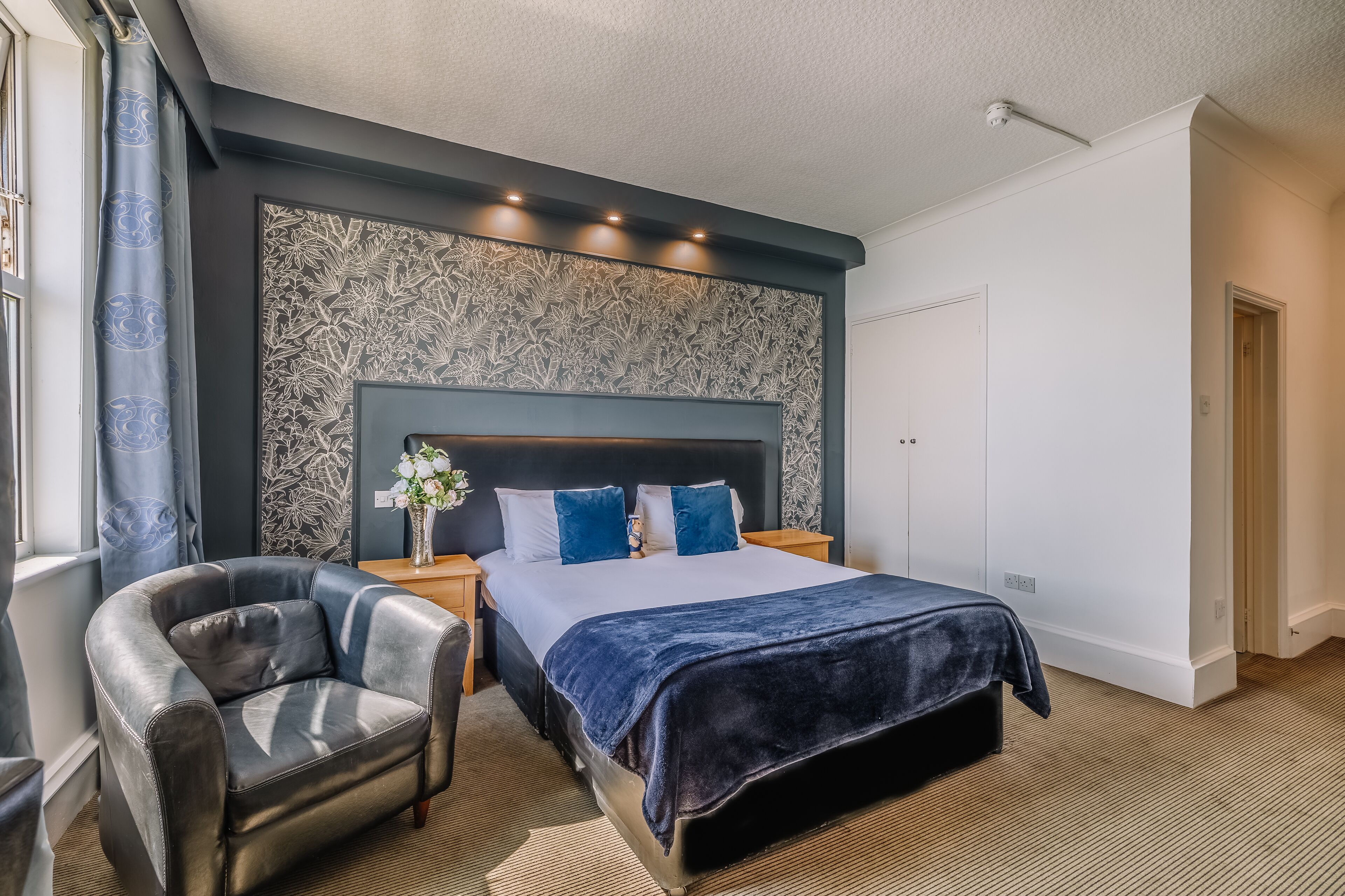 A modern bedroom with a large bed, a headboard with a floral pattern, a dark blue throw, a leather armchair, and a nightstand with a vase of flowers.