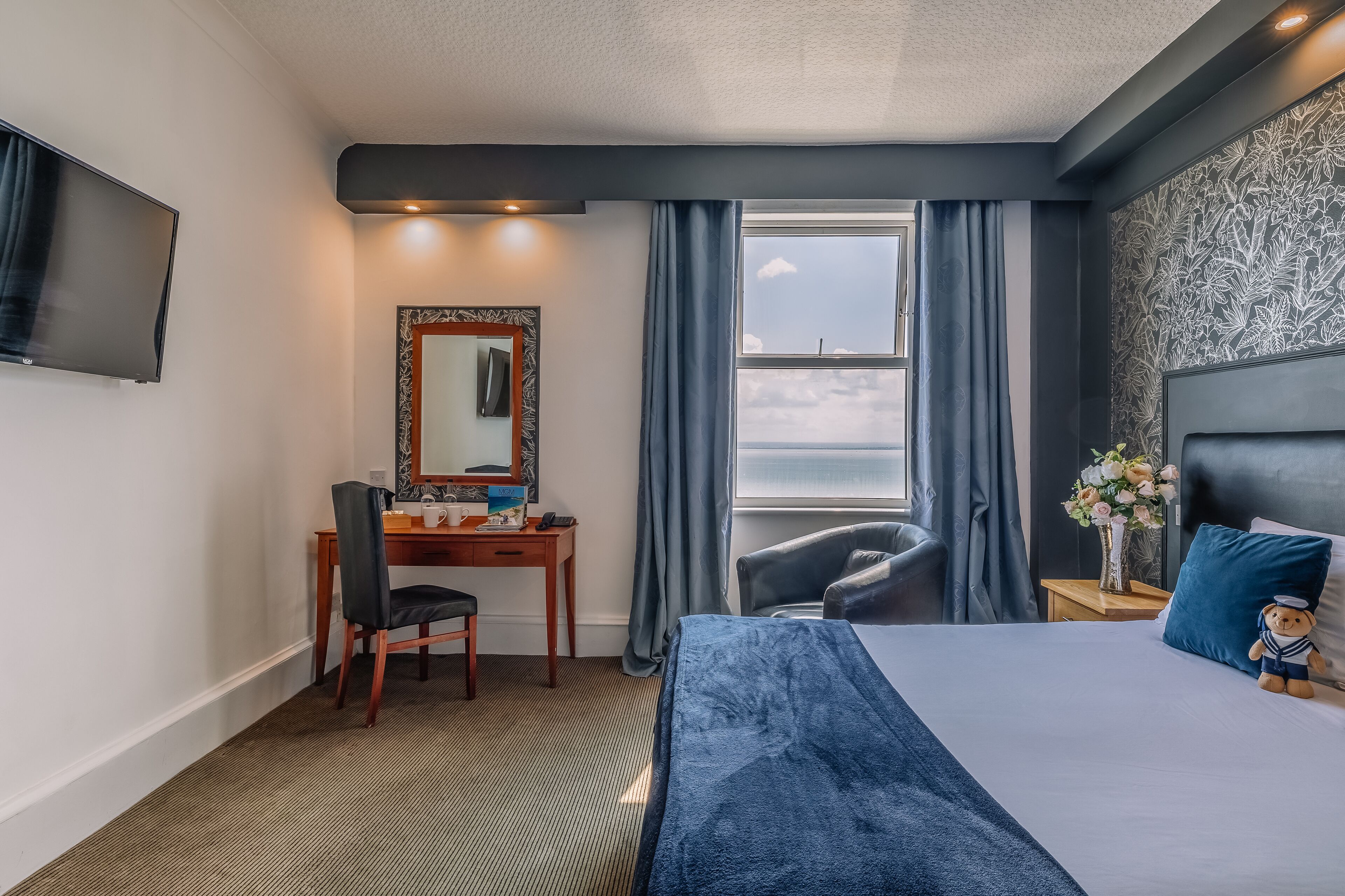 Habitación de hotel con cama, escritorio, silla, televisor, ventana con cortinas y mesita de noche con un jarrón de flores.