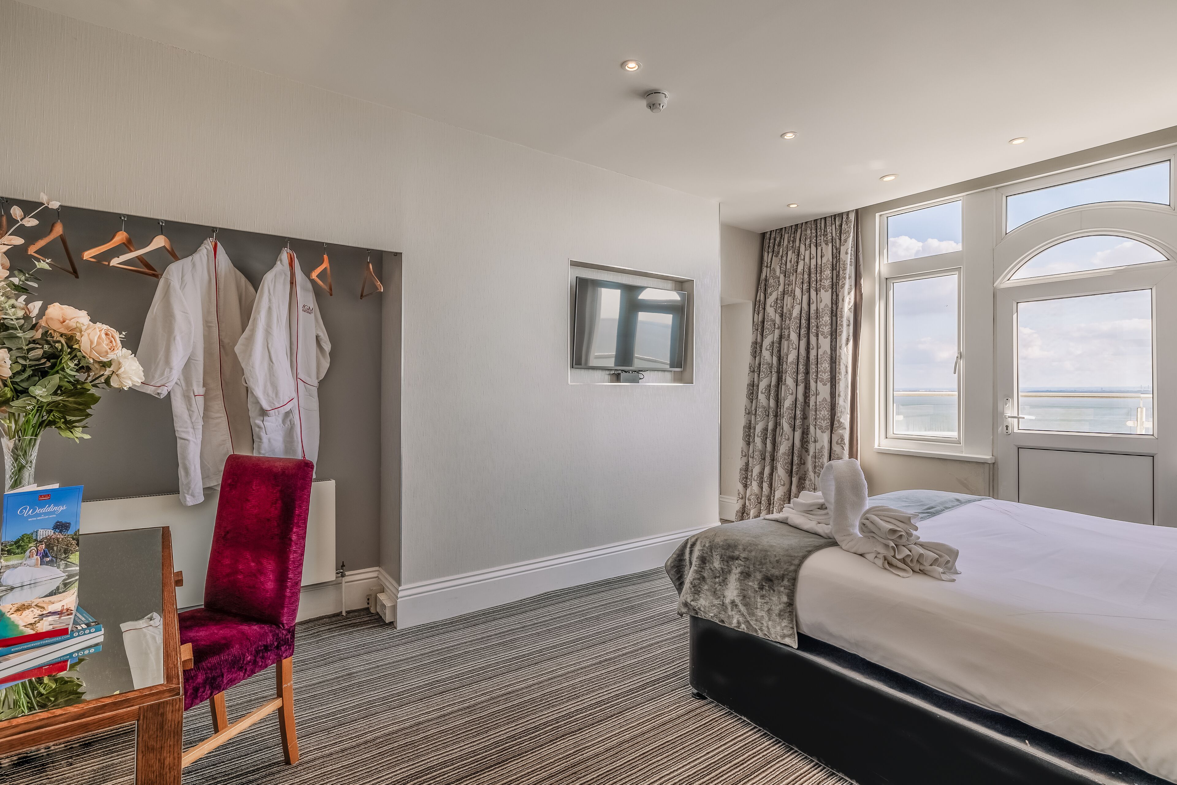 A hotel room with a bed, a desk with a chair, a window with curtains, and a view of the sky.