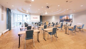 A conference room with multiple tables and chairs, a projector screen, and a large window with a view of buildings. - Scandic Vadsø (Vadsø)