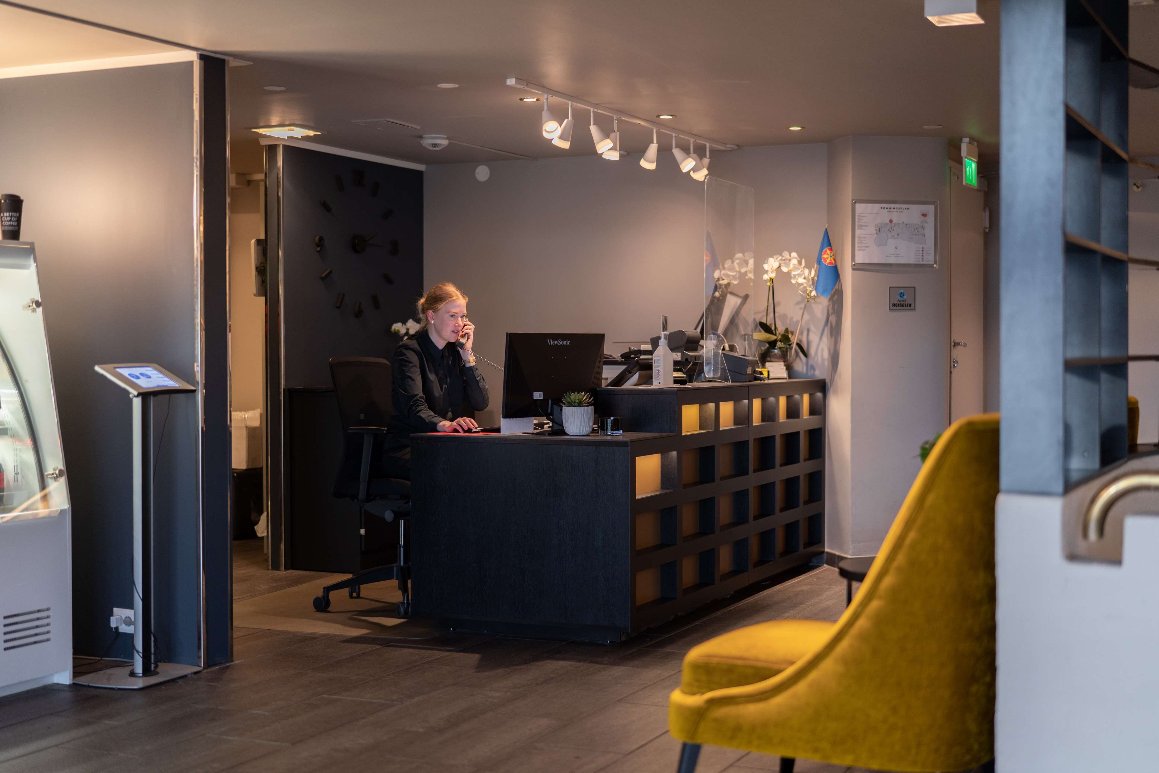 A reception desk with a computer, a plant, and a flag. There is a yellow chair in the foreground.