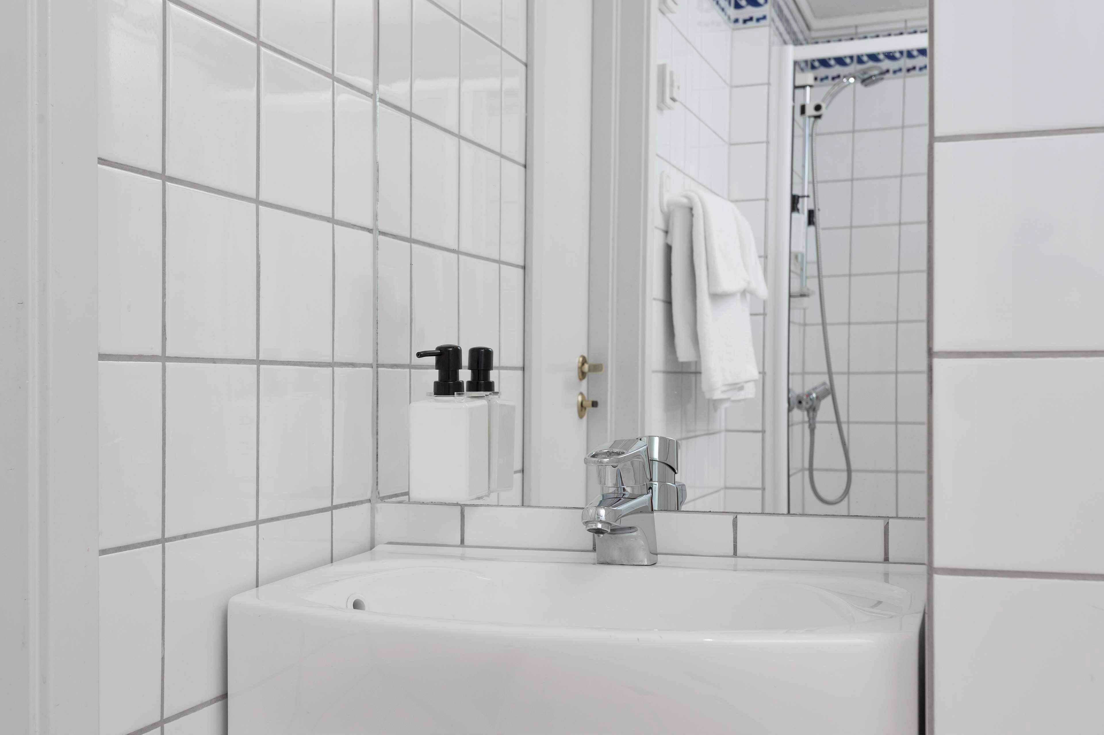 Une salle de bain moderne avec des murs recouverts de carreaux blancs, un lavabo blanc, un miroir et un espace douche.