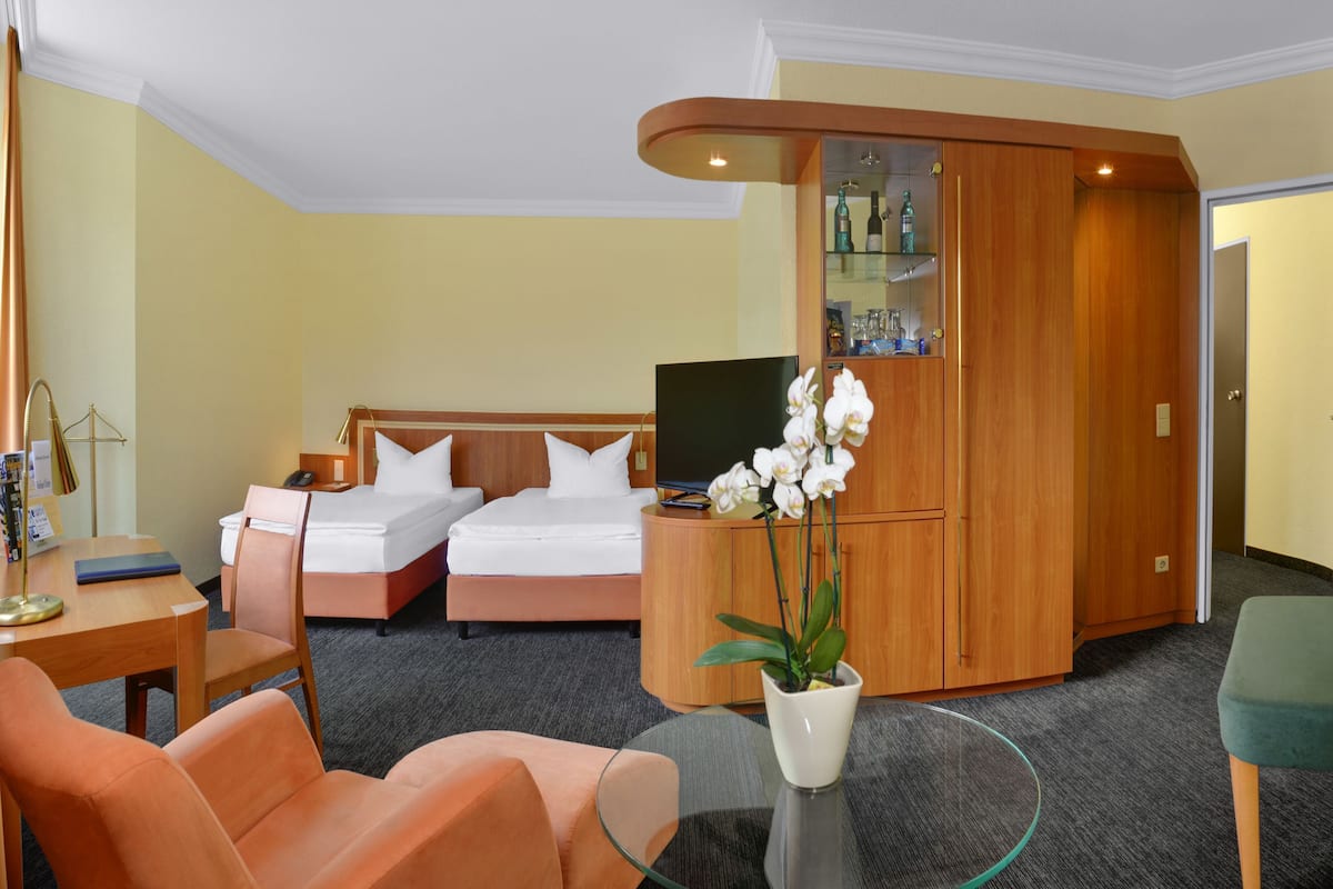 A hotel room with two beds, a desk with a lamp, a TV, a small table with a vase of flowers, and a green chair.