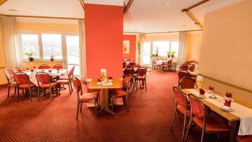 Ein Hotelrestaurant mit mehreren Tischen und Stühlen, großen Fenstern und einer roten Akzentwand.