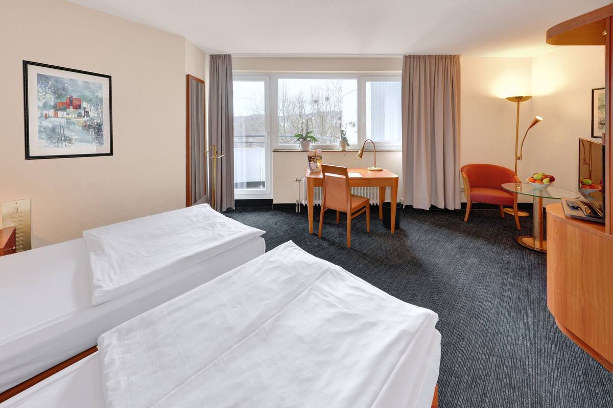 A hotel room with two beds, a desk, a chair, a lamp, and a window with curtains.