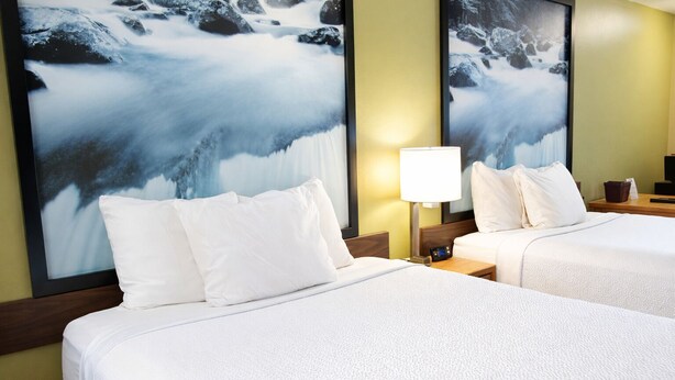 Ein Hotelzimmer mit zwei Betten, weißer Bettwäsche, einem Nachttisch mit Lampe und einem großen gerahmten Bild einer verschneiten Landschaft an der Wand.