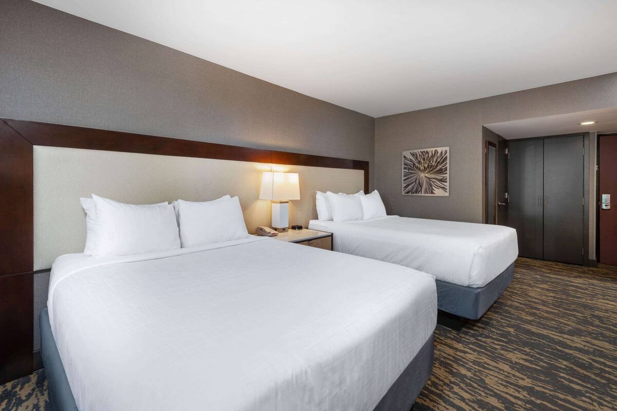 A hotel room with two beds, a nightstand with a lamp, a wall art piece, and a wardrobe.