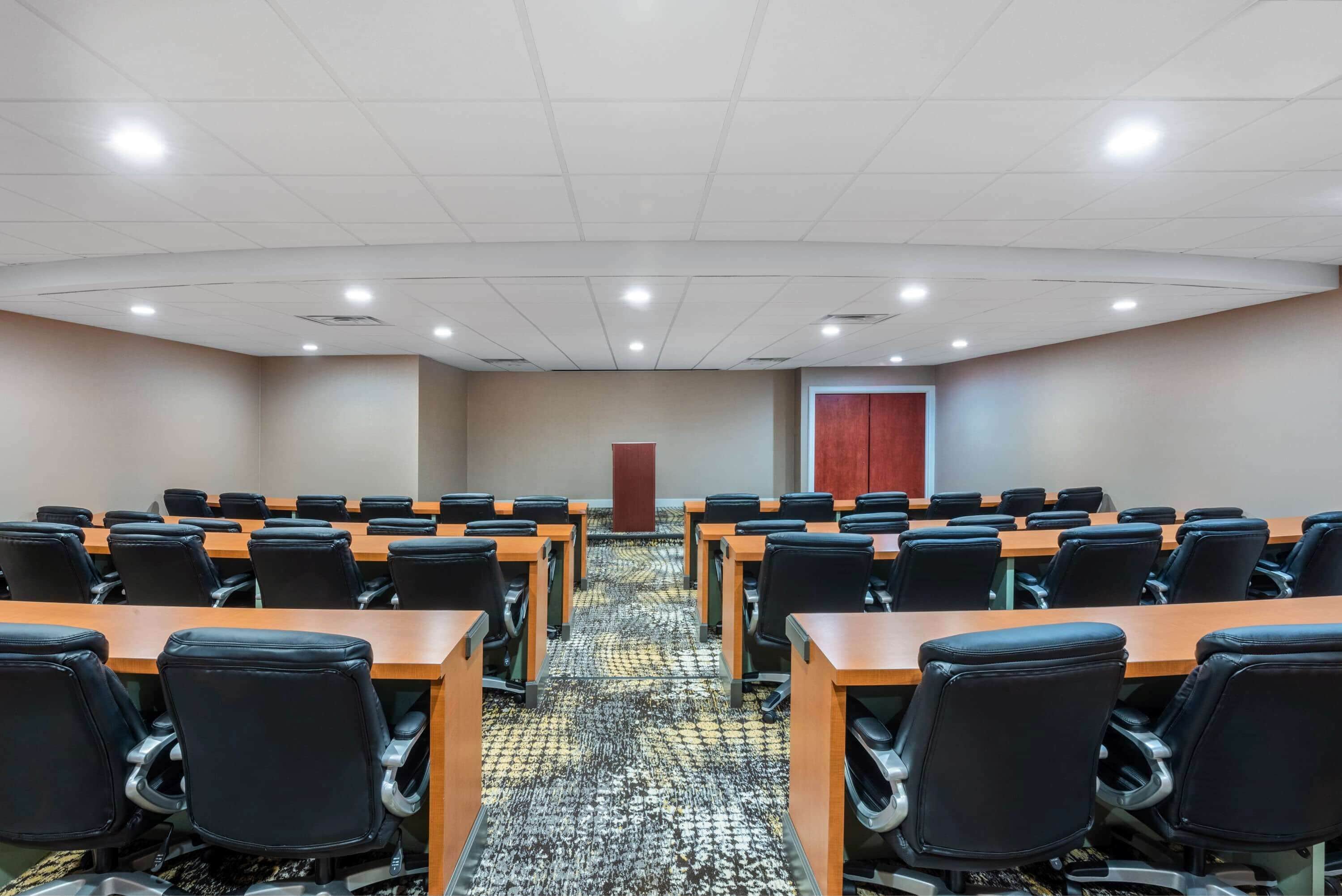 A conference room with rows of tables and chairs, a patterned carpet, and a door in the background.