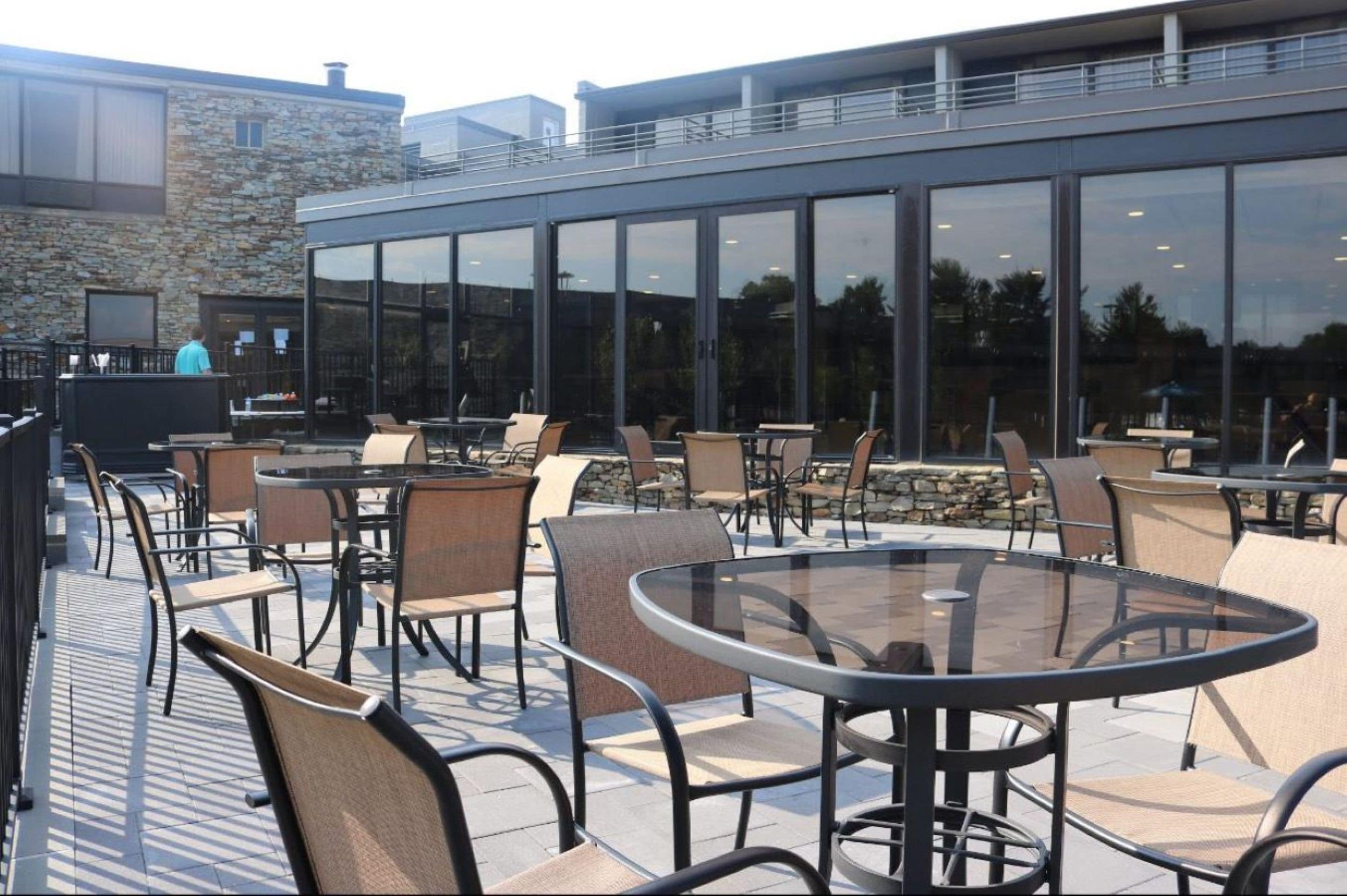 An outdoor patio with round tables and chairs, a stone wall, and a modern building with large windows.