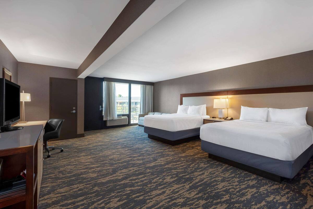 A hotel room with two beds, a desk with a TV, and a view of the outside.