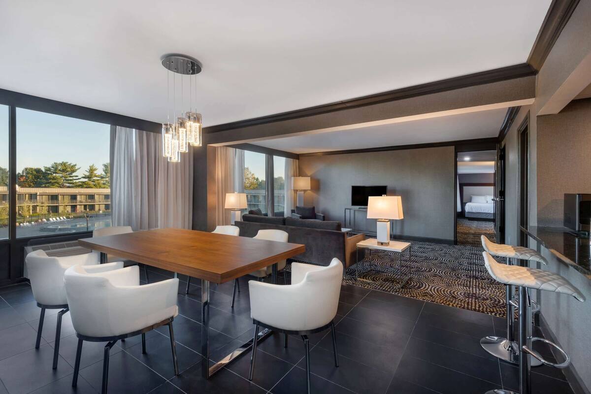 A modern hotel room with a dining area, a sofa, a TV, and a large window offering a city view.