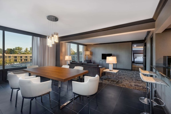 A modern hotel room with a dining area, a sofa, a TV, and a large window offering a city view.