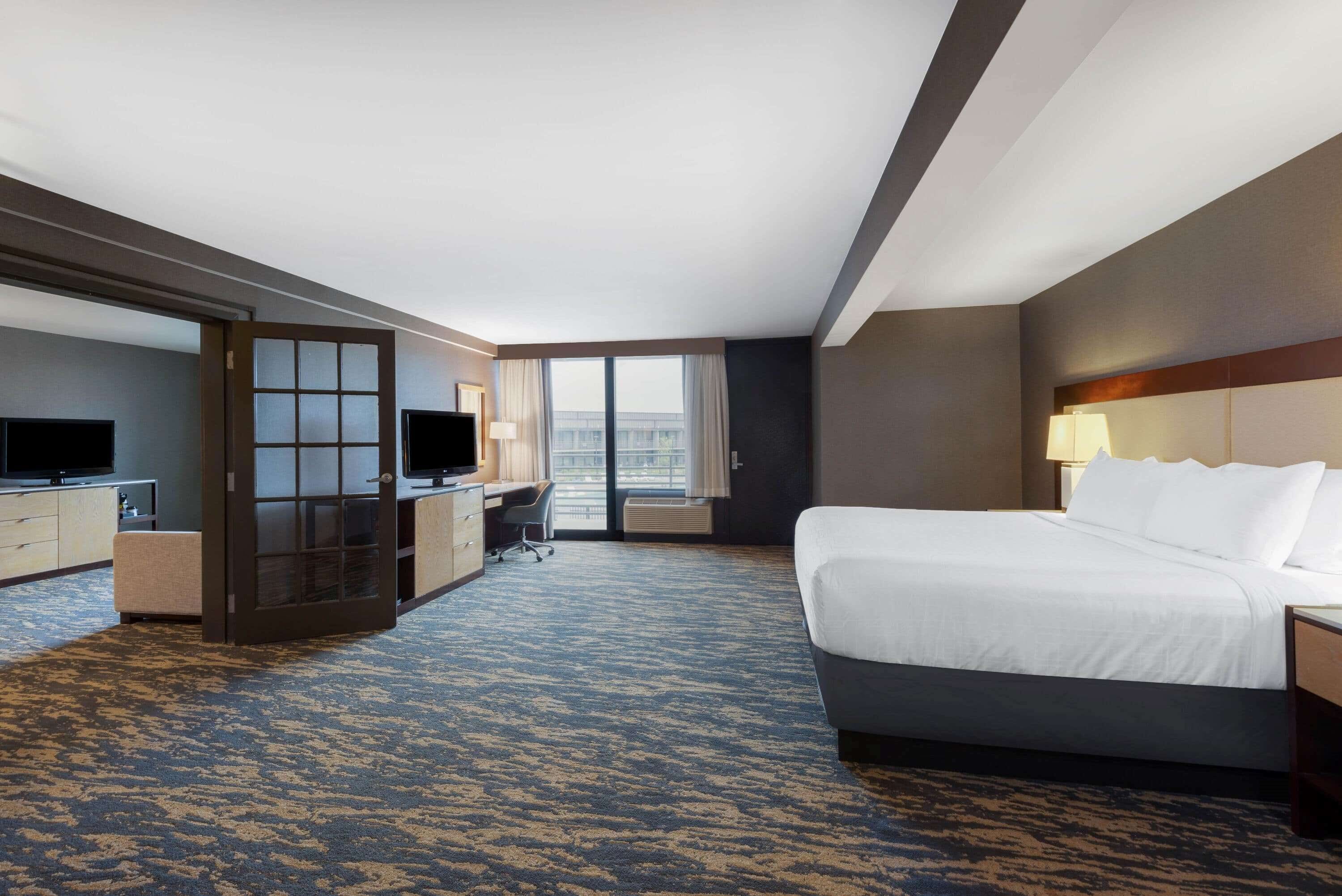 A hotel room with a bed, desk, TV, and a large window.