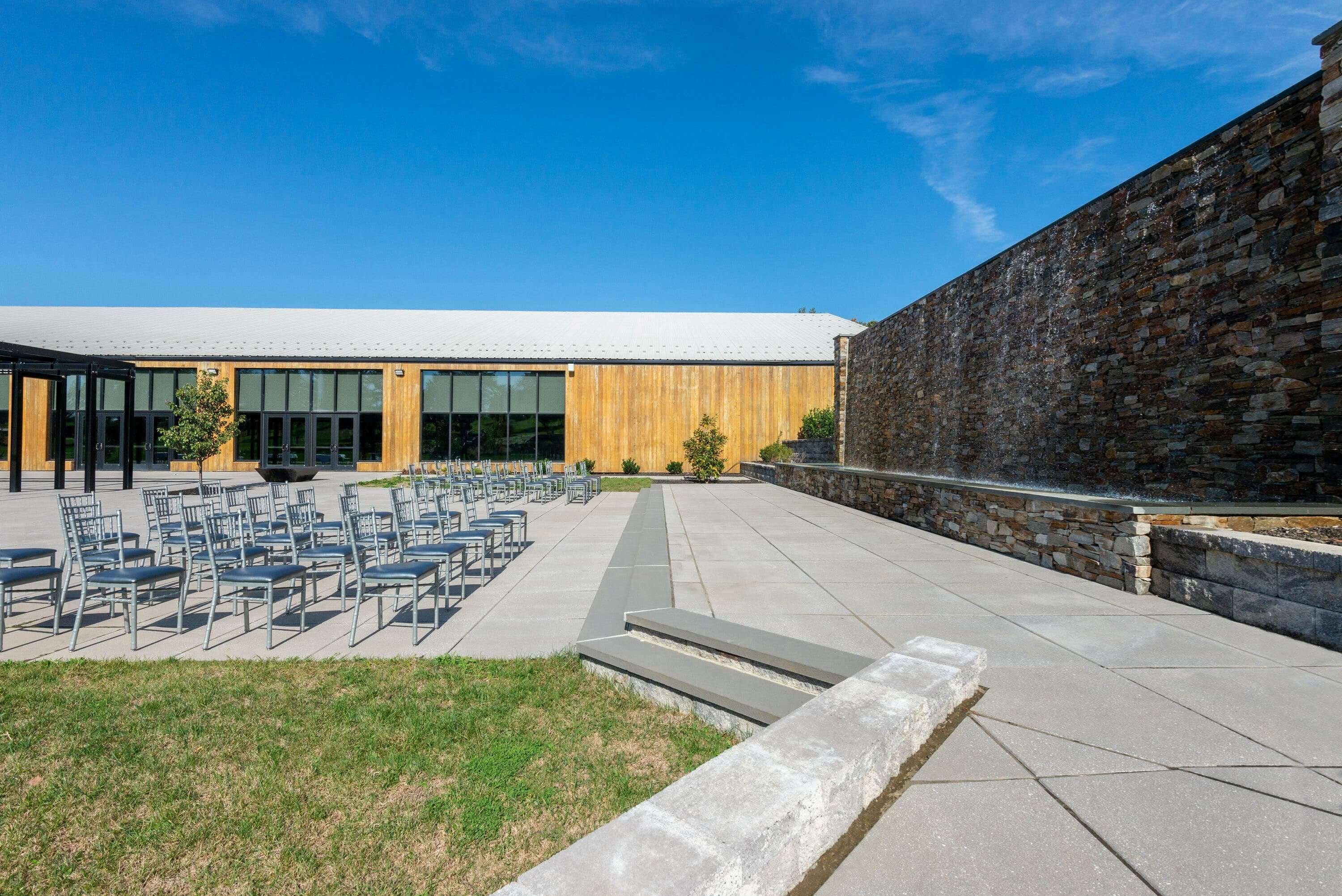 Un bâtiment moderne doté d’un mur en pierre et d’une cour dallée où sont disposées des rangées de chaises.