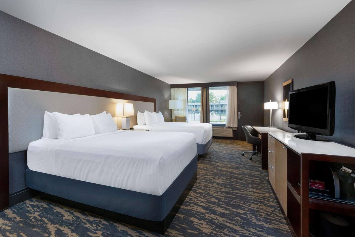 A hotel room with two beds, a desk, a television, and a view of a building through the window.