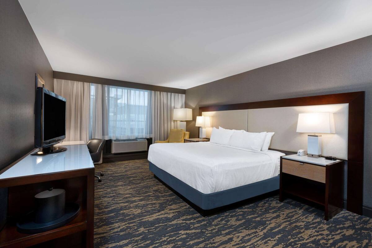 A hotel room with a large bed, a desk with a flat-screen TV, two bedside lamps, a chair, and a window with curtains.