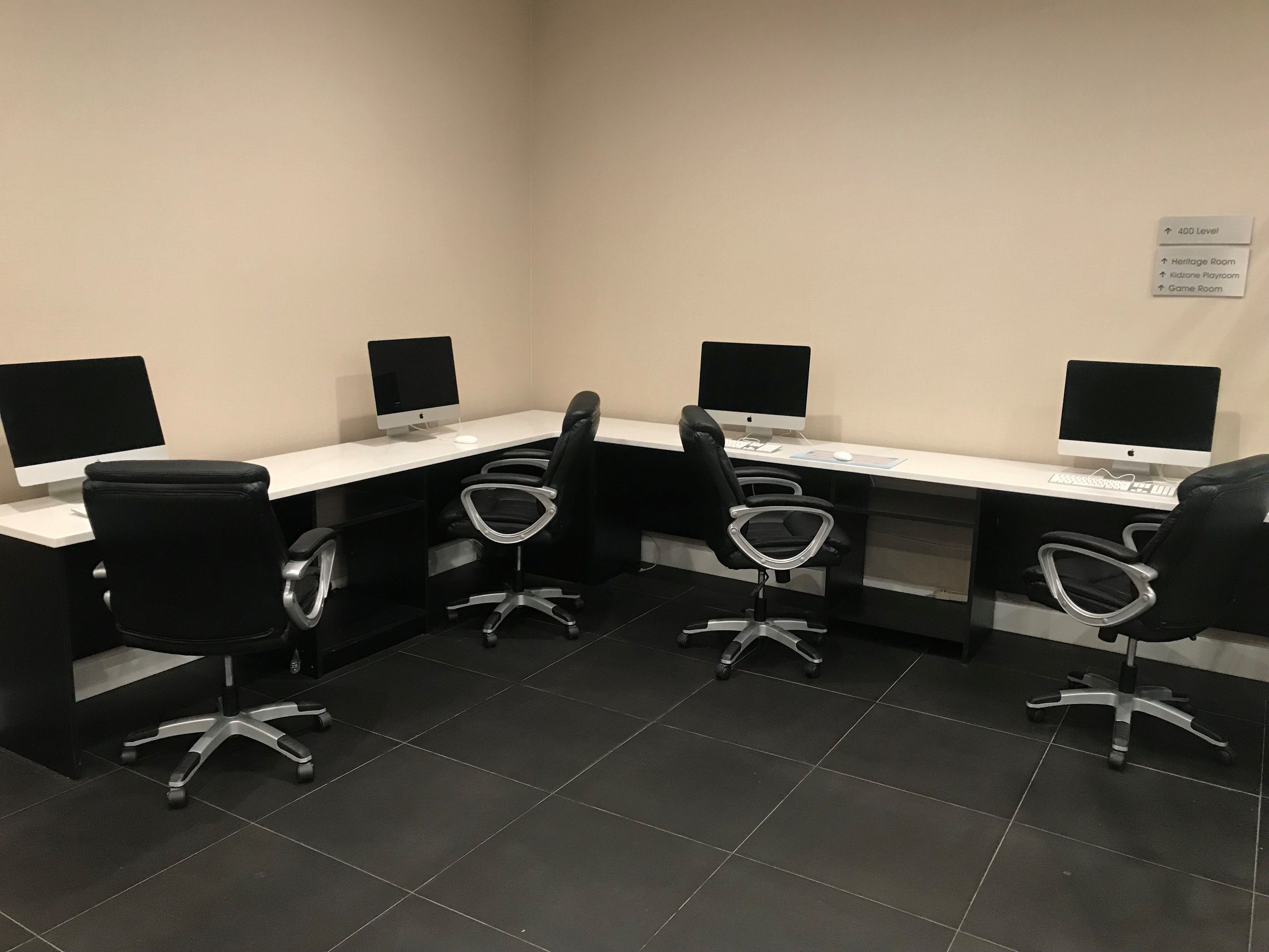 A conference room with four desks, each with a computer and a chair. There is a sign indicating the location of the Heritage Room, Kidzone Playroom, and Game Room.